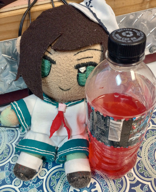 a photo of a homemade plushie of Captain Murasa from Touhou in the style of the Fumofumo line of plushies. He is being held in front of a trans flag